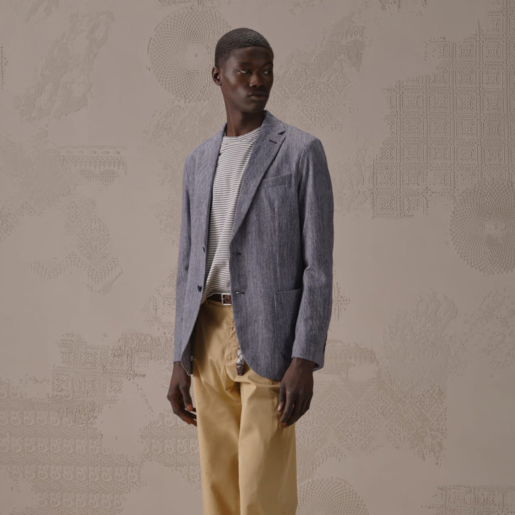 A man wearing a textured blue-gray blazer over a striped white shirt and beige trousers, standing against a beige patterned wall. Circolo 1901 Blazer. Sakks Menswear Southampton.