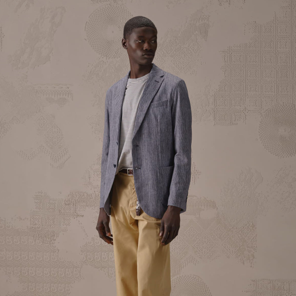 A man wearing a textured blue-gray blazer over a striped white shirt and beige trousers, standing against a beige patterned wall. Circolo 1901 Blazer. Sakks Menswear Southampton.