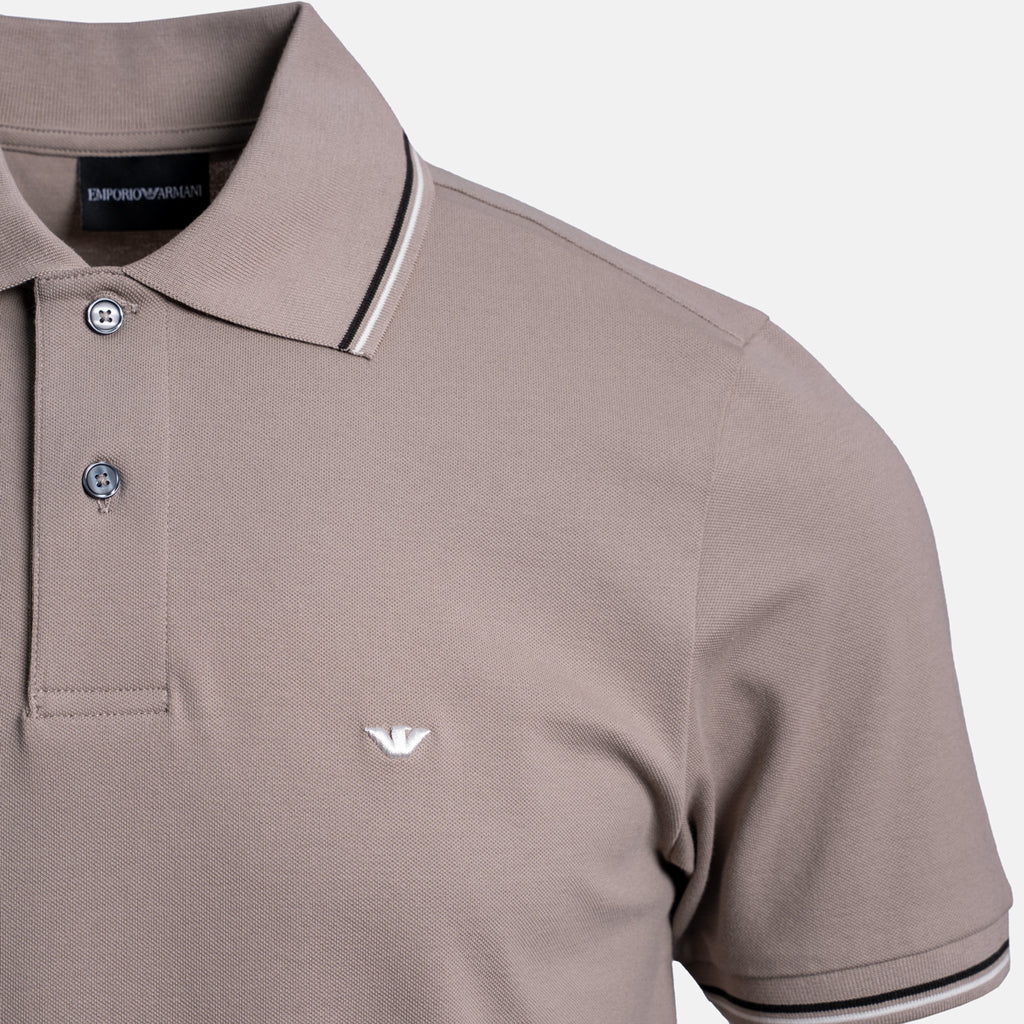 A close-up of an Emporio Armani polo shirt in taupe, featuring a white embroidered eagle logo on the chest, a two-button placket, and contrasting striped trim on the collar and sleeve cuffs. Emporio Armani Polo. Sakks Menswear Southampton.
