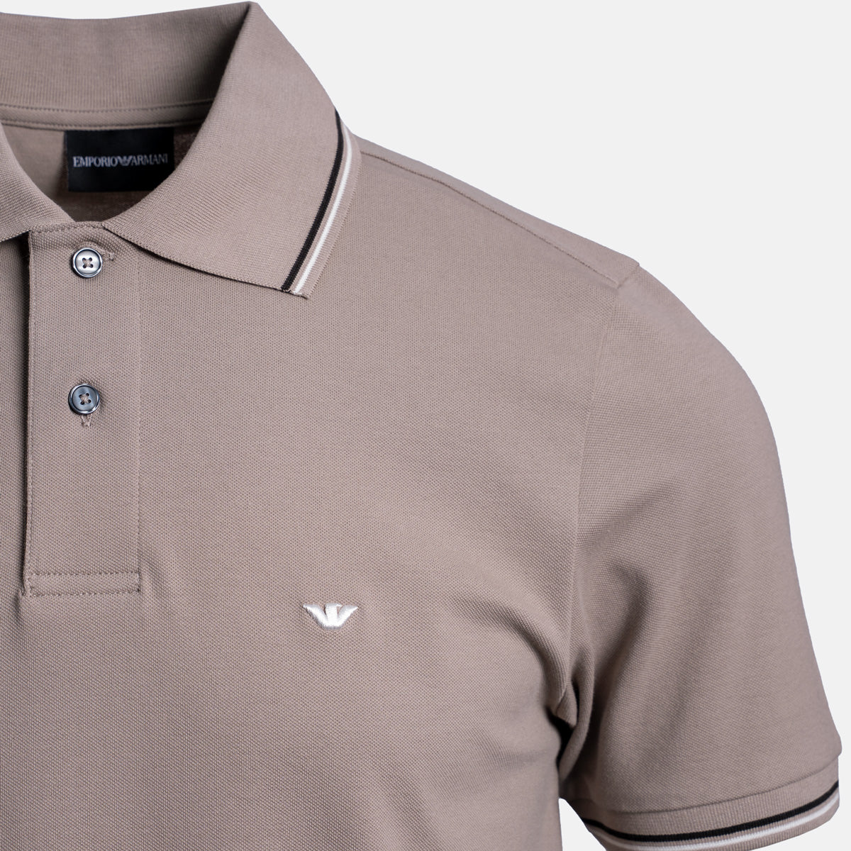 A close-up of an Emporio Armani polo shirt in taupe, featuring a white embroidered eagle logo on the chest, a two-button placket, and contrasting striped trim on the collar and sleeve cuffs. Emporio Armani Polo. Sakks Menswear Southampton.