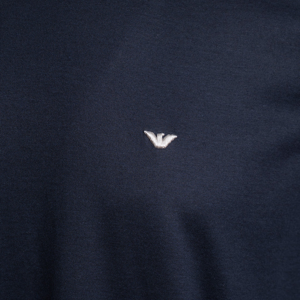 Close-up of a dark navy blue knit fabric with a small white embroidered eagle logo at the center. Emporio Armani T-Shirt. Sakks Menswear Southampton.