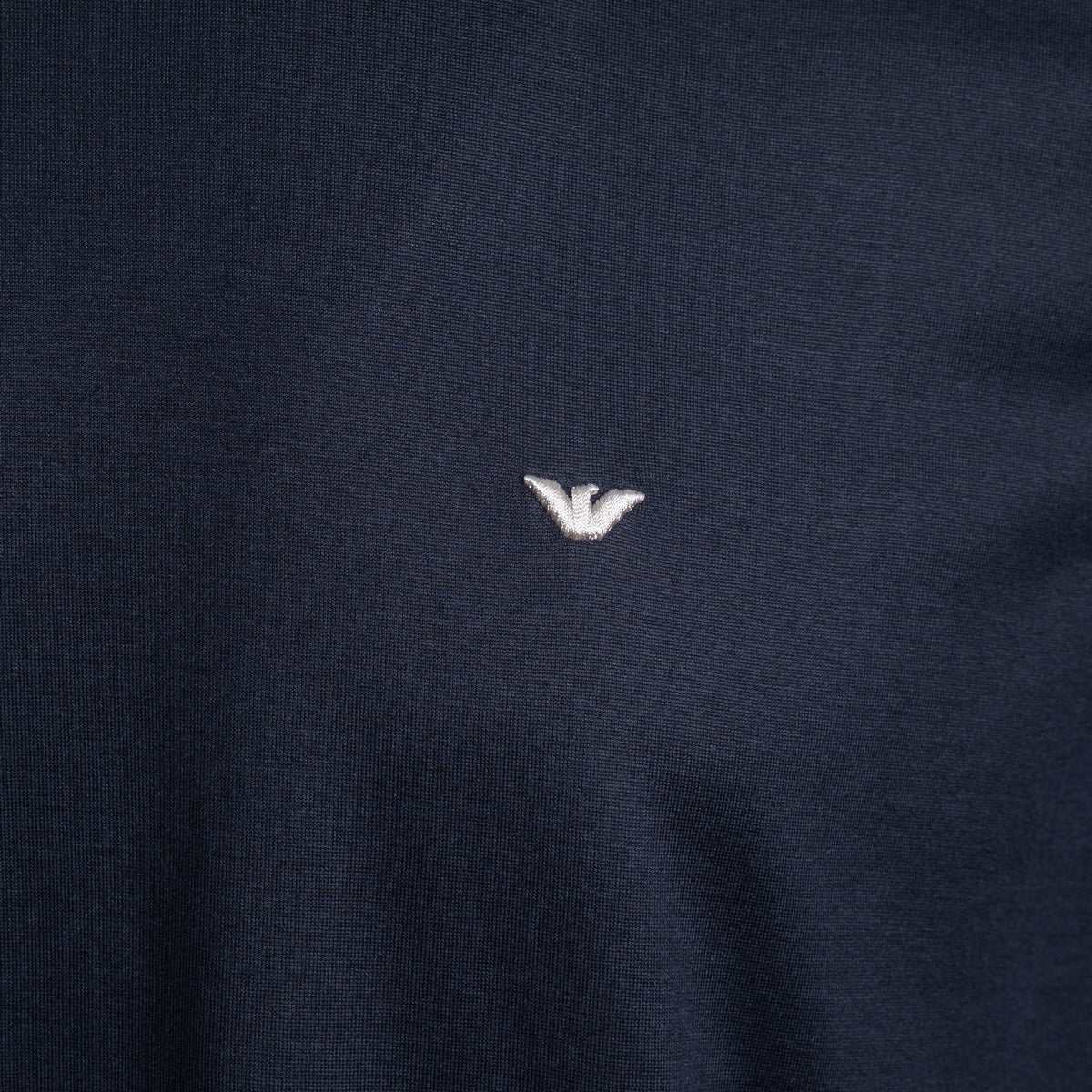 Close-up of a dark navy blue knit fabric with a small white embroidered eagle logo at the center. Emporio Armani T-Shirt. Sakks Menswear Southampton.