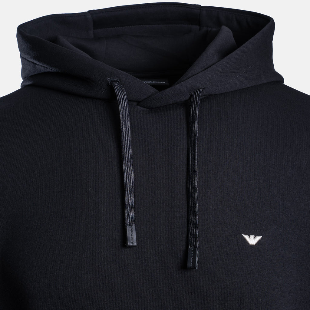 Close-up of a black pullover hoodie with matching black drawstrings and a small white embroidered eagle logo on the left chest, shown against a light gray background. Emporio Armani Hoodie. Sakks Menswear Southampton.