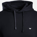 Close-up of a black pullover hoodie with matching black drawstrings and a small white embroidered eagle logo on the left chest, shown against a light gray background. Emporio Armani Hoodie. Sakks Menswear Southampton.