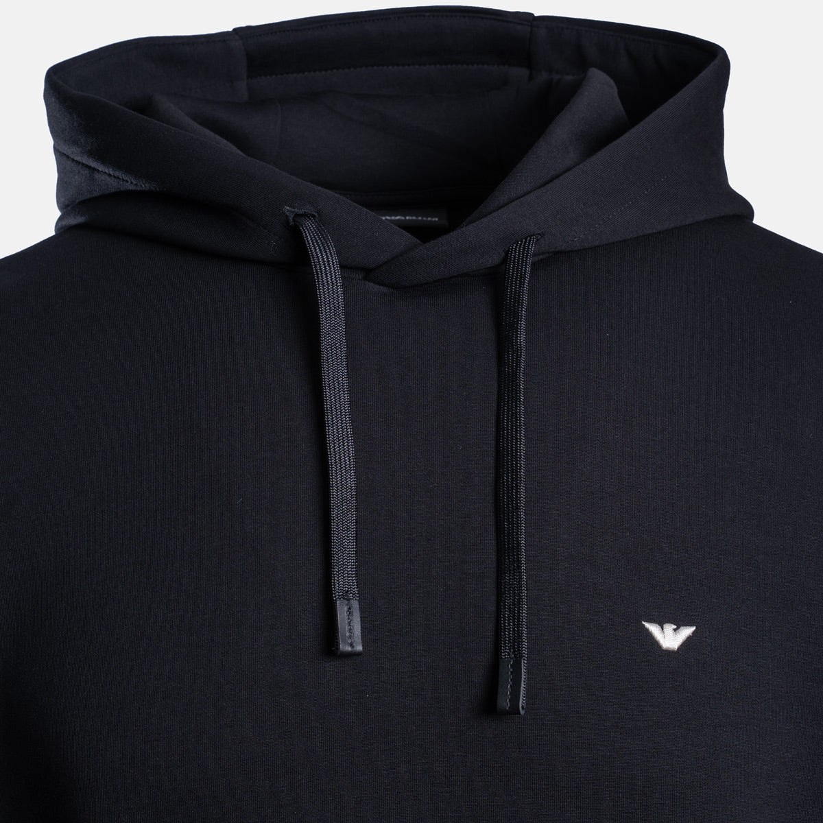Close-up of a black pullover hoodie with matching black drawstrings and a small white embroidered eagle logo on the left chest, shown against a light gray background. Emporio Armani Hoodie. Sakks Menswear Southampton.