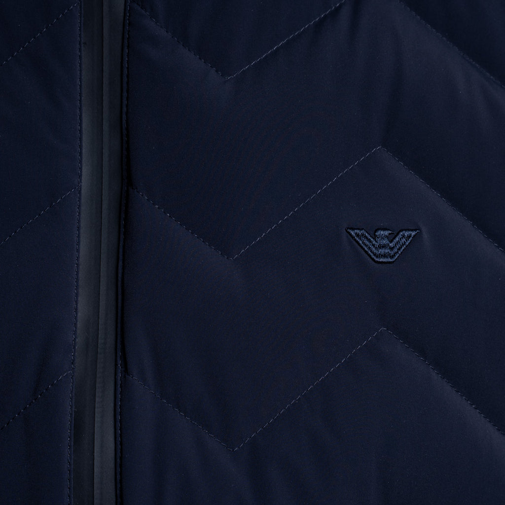 Close-up of a navy blue quilted jacket with a chevron pattern, a black zipper, and an embroidered dark logo on the chest. Emporio Armani Gilet. Sakks Menswear Southampton.