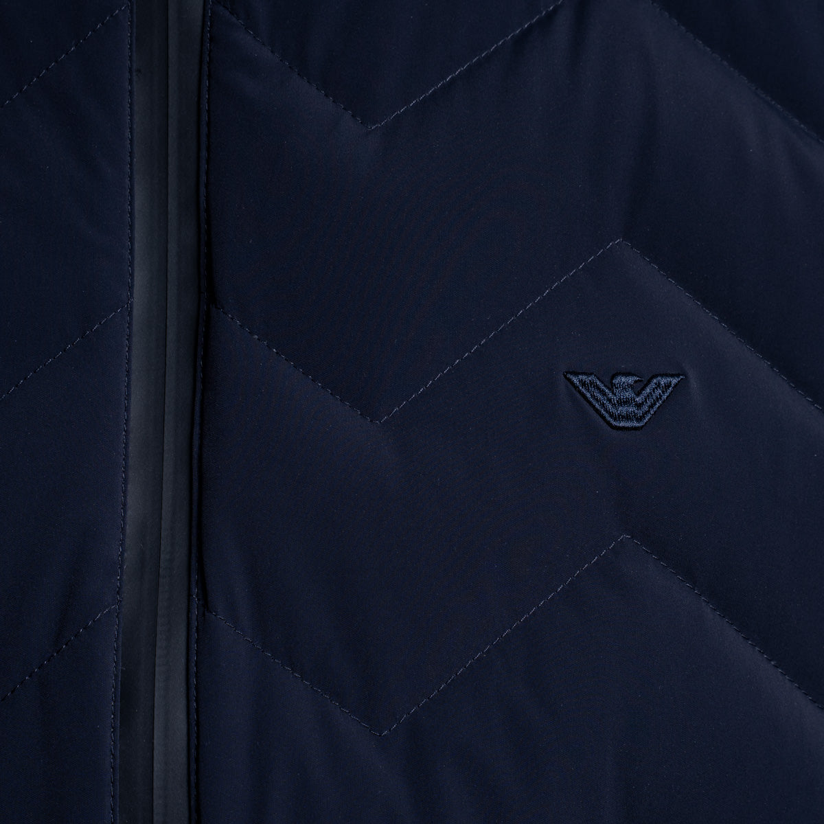 Close-up of a navy blue quilted jacket with a chevron pattern, a black zipper, and an embroidered dark logo on the chest. Emporio Armani Gilet. Sakks Menswear Southampton.