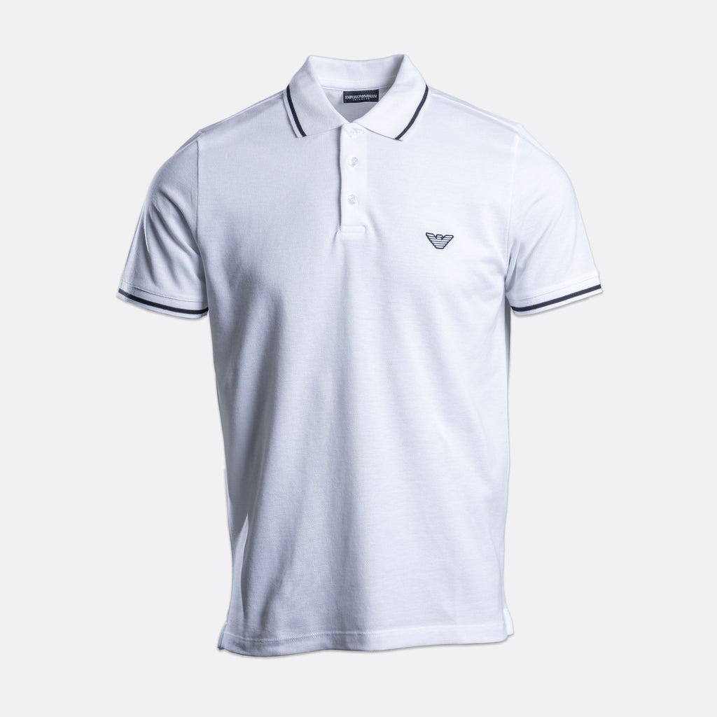 A white short-sleeve polo shirt with navy blue trim on the collar and sleeve cuffs, featuring a small embroidered logo on the left chest, displayed on a light gray background. Emporio Armani Polo. Sakks Menswear Southampton.