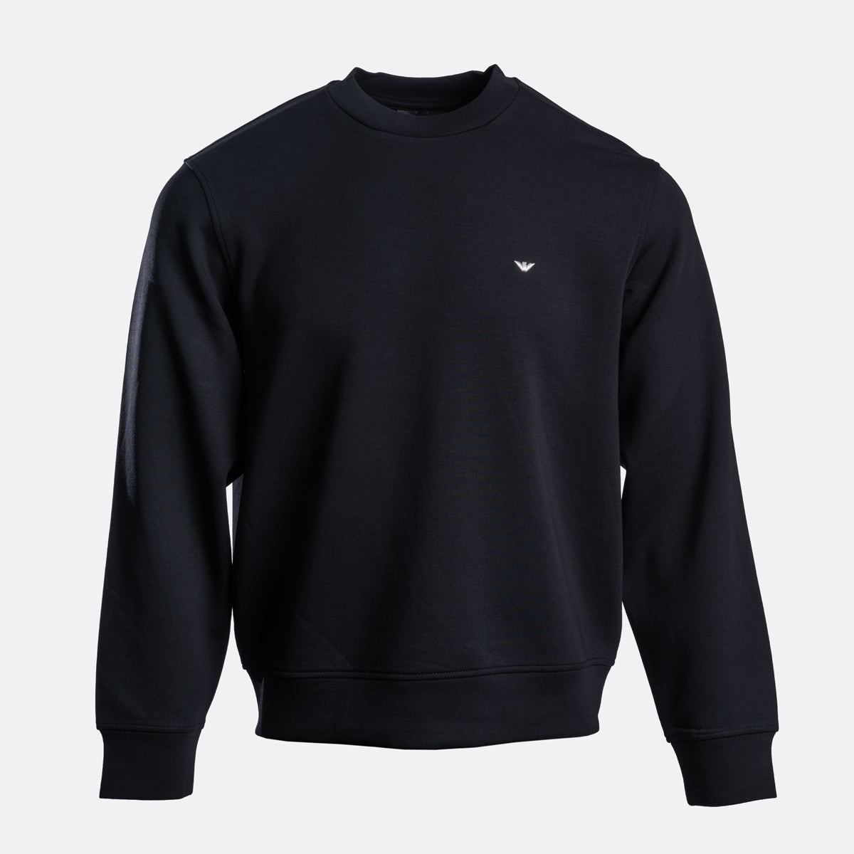 A black crewneck sweatshirt with a small white embroidered eagle logo on the left chest, displayed on a clean light gray background. Emporio Armani Sweatshirt. Sakks Menswear Southampton.