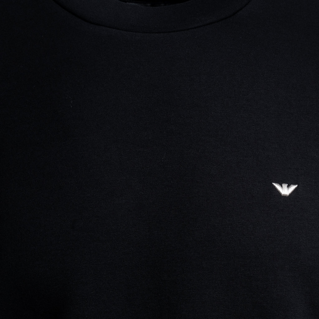 Close-up of a black crewneck sweatshirt featuring a small white embroidered eagle logo on the left chest, representing the brand Emporio Armani. Emporio Armani Sweatshirt. Sakks Menswear Southampton.