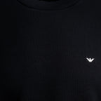 Close-up of a black crewneck sweatshirt featuring a small white embroidered eagle logo on the left chest, representing the brand Emporio Armani. Emporio Armani Sweatshirt. Sakks Menswear Southampton.