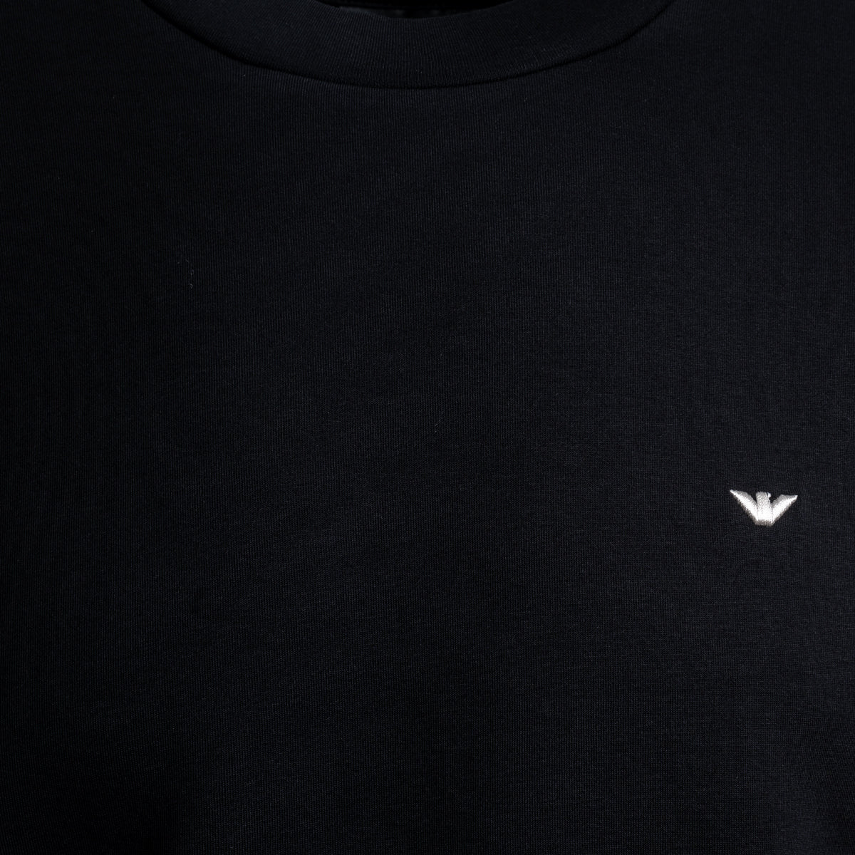 Close-up of a black crewneck sweatshirt featuring a small white embroidered eagle logo on the left chest, representing the brand Emporio Armani. Emporio Armani Sweatshirt. Sakks Menswear Southampton.