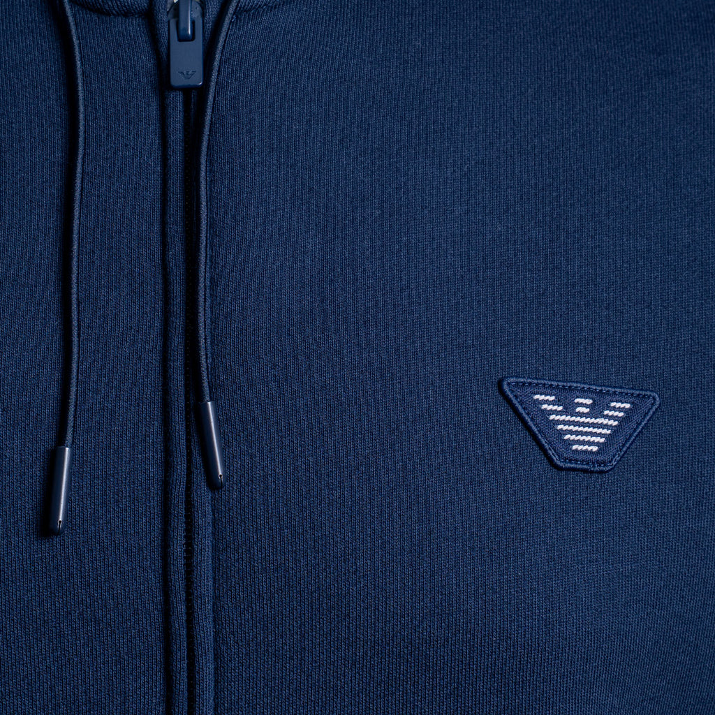 Close-up of a navy blue zip-up hoodie showing the front zipper, drawstrings, and a small embroidered triangular logo patch on the chest. Emporio Armani Hoodie. Sakks Menswear Southampton.