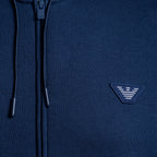 Close-up of a navy blue zip-up hoodie showing the front zipper, drawstrings, and a small embroidered triangular logo patch on the chest. Emporio Armani Hoodie. Sakks Menswear Southampton.