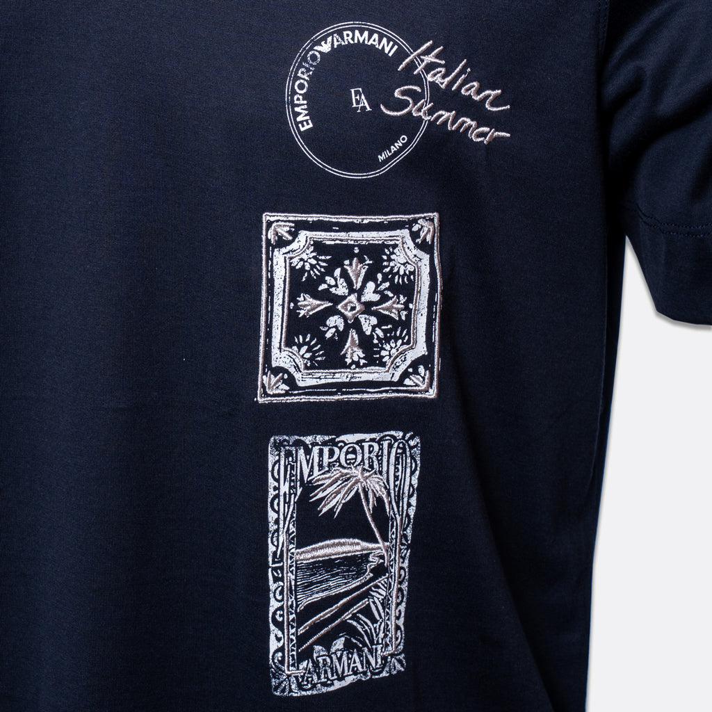 Close-up of a navy blue t-shirt featuring three vertical white graphic prints. The top graphic is a circular stamp reading 'EMPORIO ARMANI Milano'.