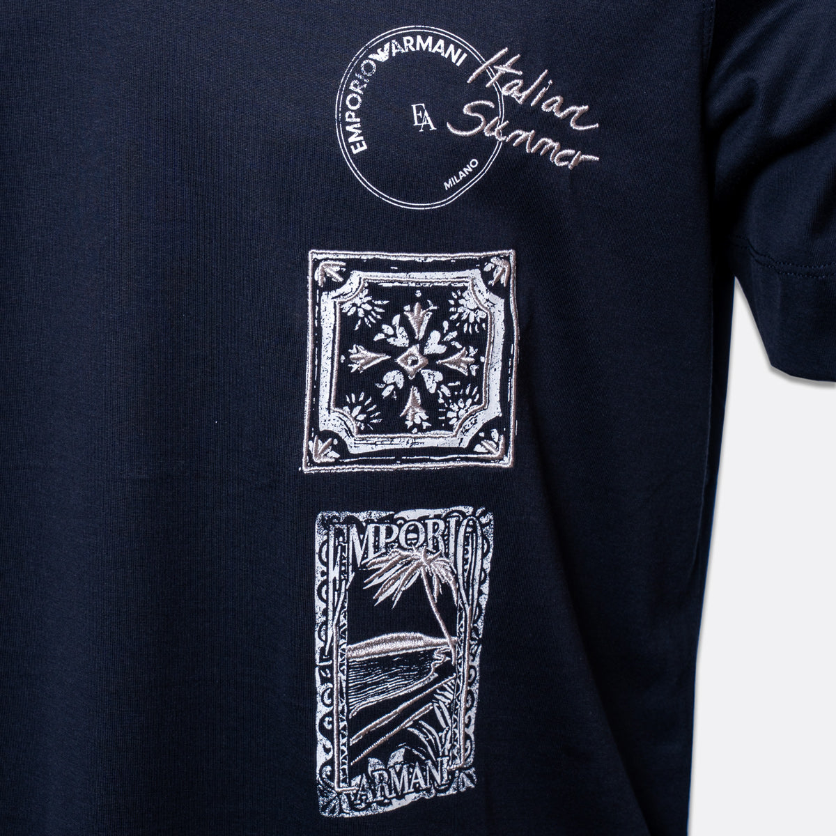 Close-up of a navy blue t-shirt featuring three vertical white graphic prints. The top graphic is a circular stamp reading 'EMPORIO ARMANI Milano'.