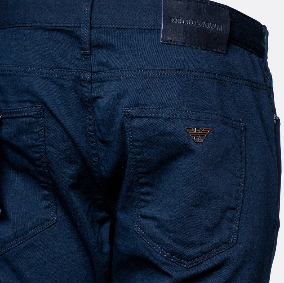 Close-up of the back waistband and right rear pocket of dark navy blue jeans, showing a black leather patch with 'EMPORIO ARMANI' text and a small embroidered logo on the pocket. Emporio Armani Chinos. Sakks Menswear Southampton.