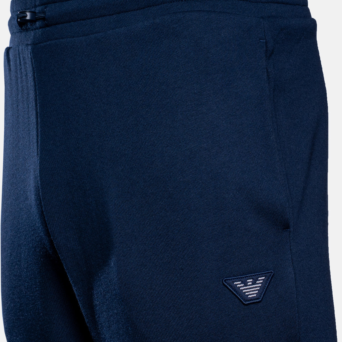 Close-up of navy blue sweatpants showing the fabric texture, side pocket seam, and a small embroidered triangular logo patch on the lower leg. Emporio Armani Sweat Pants. Sakks Menswear Southampton.