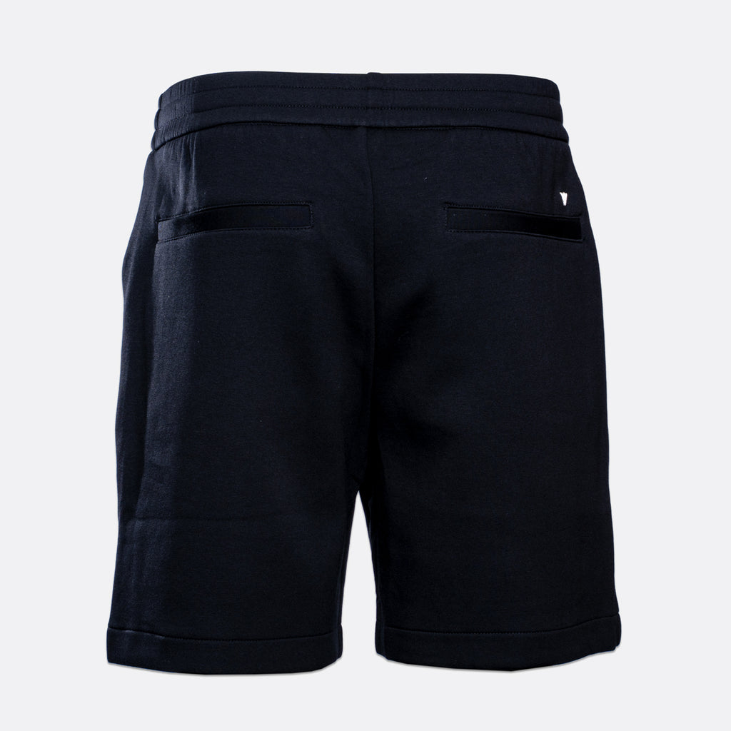 Rear view of dark navy sweat shorts with an elastic waistband, side pockets, and a small white embroidered logo on the back right pocket, shown on a light gray background. Emporio Armani Shorts. Sakk Menswear Southampton.