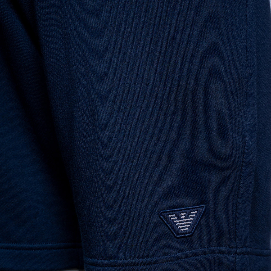 Close-up of a navy blue knit garment, likely a sweatshirt or sweater, showing the fabric texture and a small embroidered triangular logo patch near the hem. Emporio Armani Shorts. Sakks Menswear Southampton.
