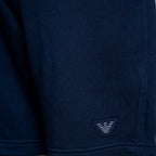 Close-up of a navy blue knit garment, likely a sweatshirt or sweater, showing the fabric texture and a small embroidered triangular logo patch near the hem. Emporio Armani Shorts. Sakks Menswear Southampton.
