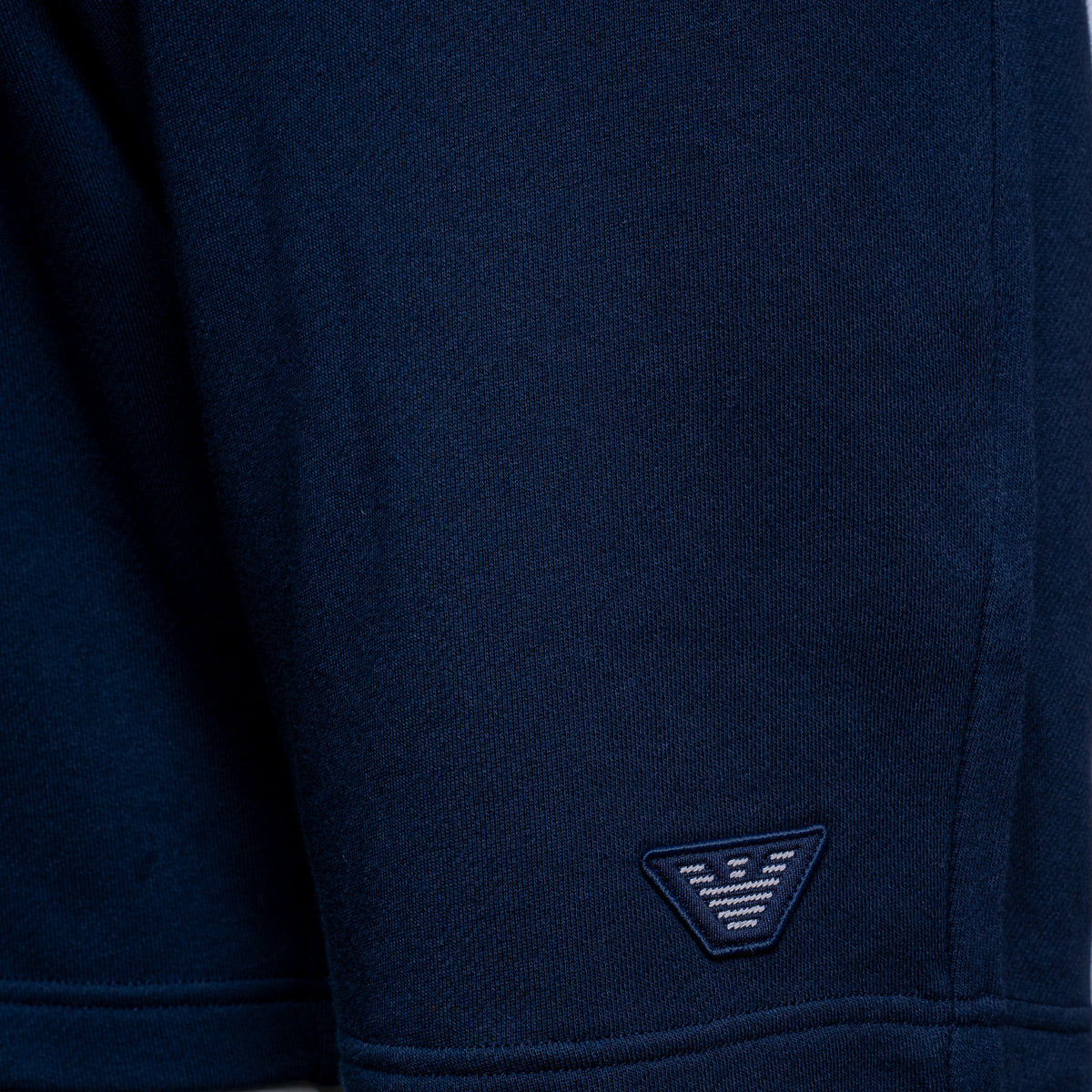 Close-up of a navy blue knit garment, likely a sweatshirt or sweater, showing the fabric texture and a small embroidered triangular logo patch near the hem. Emporio Armani Shorts. Sakks Menswear Southampton.