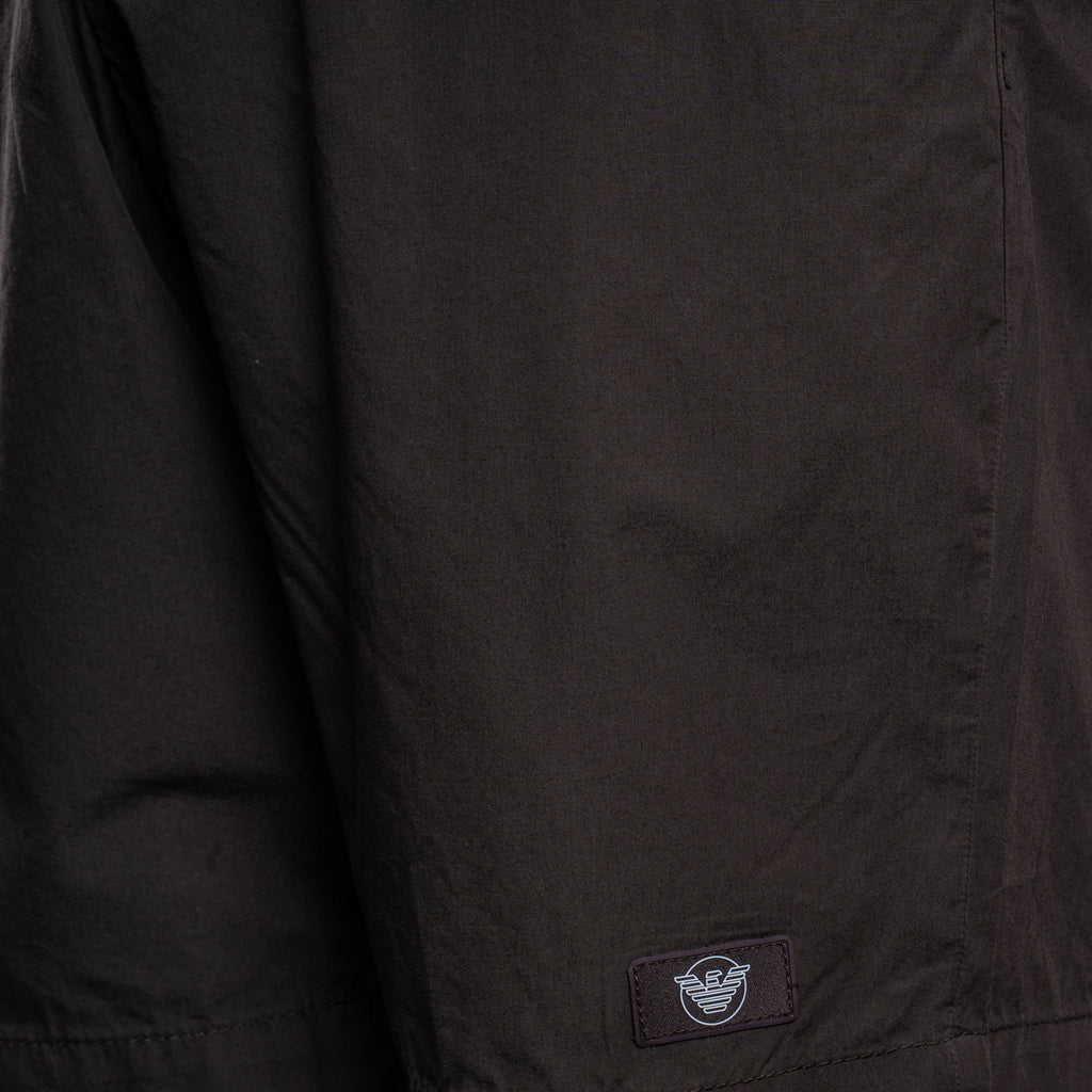 Close-up of a black lightweight jacket or overshirt fabric with a small rectangular brand patch near the lower hem. The patch shows the Armani Exchange eagle logo. Emporio Armani Shorts. Sakks Menswear Southampton.