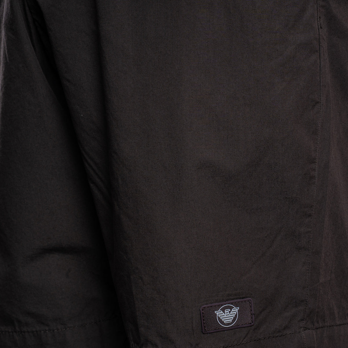 Close-up of a black lightweight jacket or overshirt fabric with a small rectangular brand patch near the lower hem. The patch shows the Armani Exchange eagle logo. Emporio Armani Shorts. Sakks Menswear Southampton.