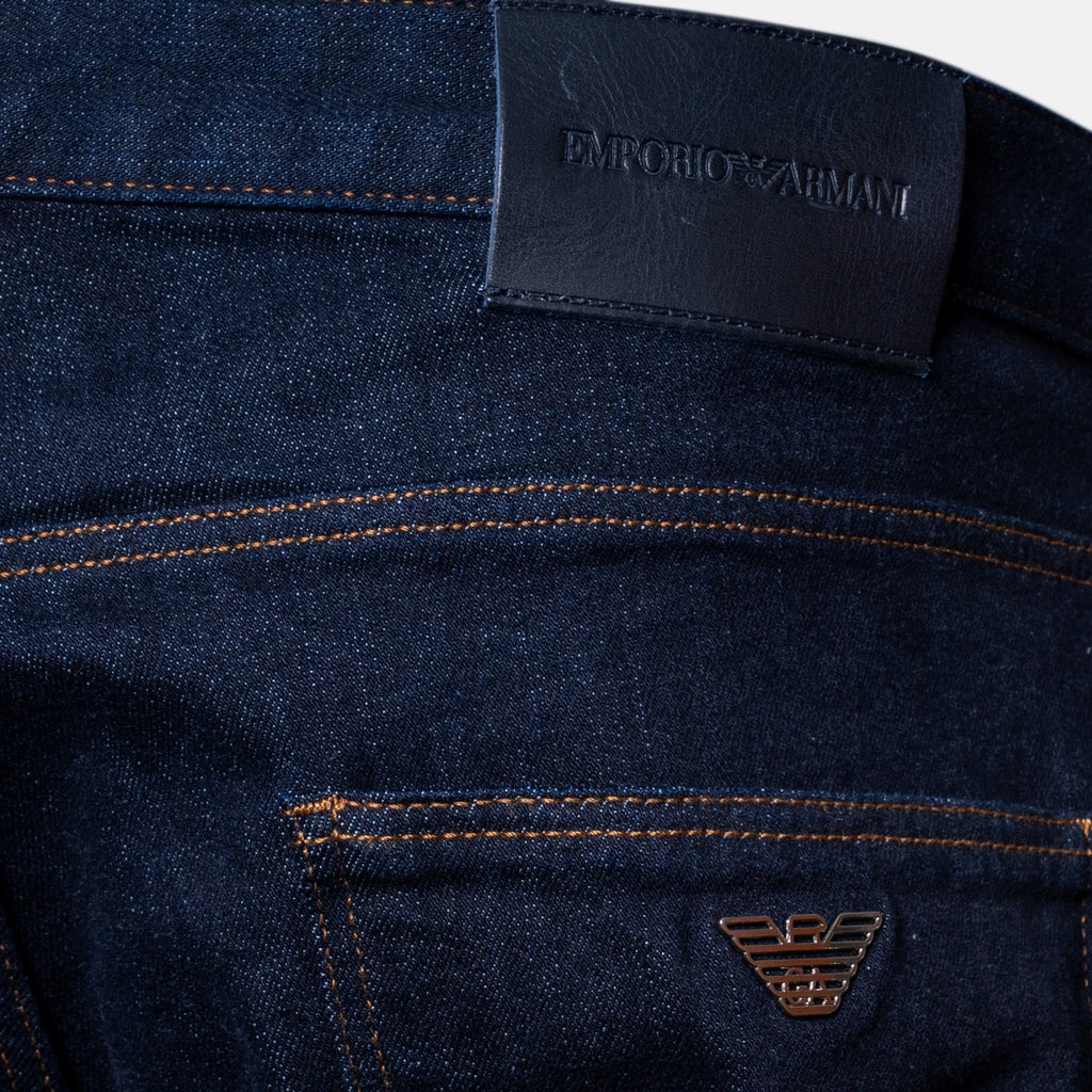 Close-up of dark indigo denim jeans showing the back waistband with a black leather patch embossed with 'EMPORIO ARMANI' and a small metallic eagle logo on the pocket. Dark blue slim-fit denim jeans shown on a light gray background. Emporio Armani Jeans. Sakks Menswear Southampton.