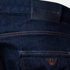 Close-up of dark indigo denim jeans showing the back waistband with a black leather patch embossed with 'EMPORIO ARMANI' and a small metallic eagle logo on the pocket. Dark blue slim-fit denim jeans shown on a light gray background. Emporio Armani Jeans. Sakks Menswear Southampton.