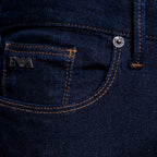 Close-up of a dark indigo denim pair of jeans showing the back pocket with orange contrast stitching, a silver metal rivet, and a silver 'EA' logo patch. Dark blue slim-fit denim jeans shown on a light gray background. Emporio Armani Jeans. Sakks Menswear Southampton.