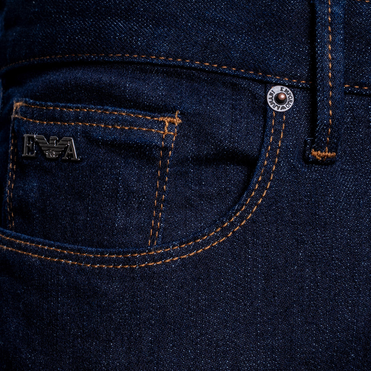 Close-up of a dark indigo denim pair of jeans showing the back pocket with orange contrast stitching, a silver metal rivet, and a silver 'EA' logo patch. Dark blue slim-fit denim jeans shown on a light gray background. Emporio Armani Jeans. Sakks Menswear Southampton.