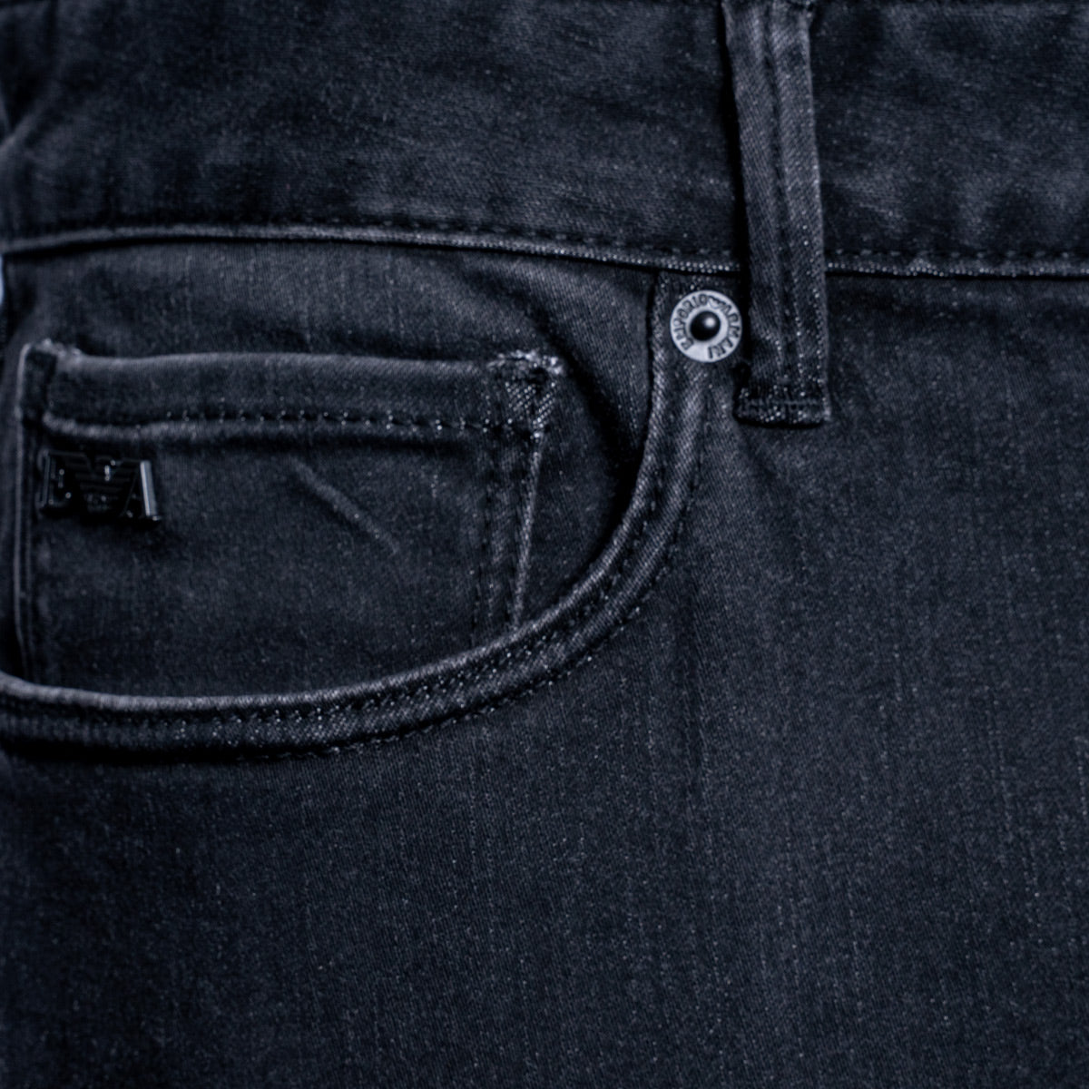 Close-up of the back pocket and waistband of dark washed denim jeans, showing a silver metal button and a small black brand tab. Emporio Armani Jeans. Sakks Menswear Southampton.