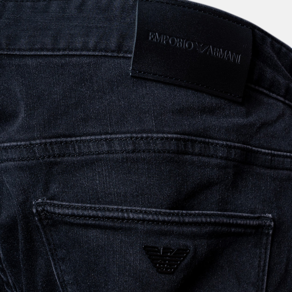 Close-up of the back waistband and pocket area of dark washed denim jeans, showing an embroidered black leather patch with the 'EMPORIO ARMANI' brand name and the signature eagle logo. Emporio Armani Jeans. Sakks Menswear Southampton.