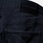 Close-up of the back waistband and pocket area of dark washed denim jeans, showing an embroidered black leather patch with the 'EMPORIO ARMANI' brand name and the signature eagle logo. Emporio Armani Jeans. Sakks Menswear Southampton.