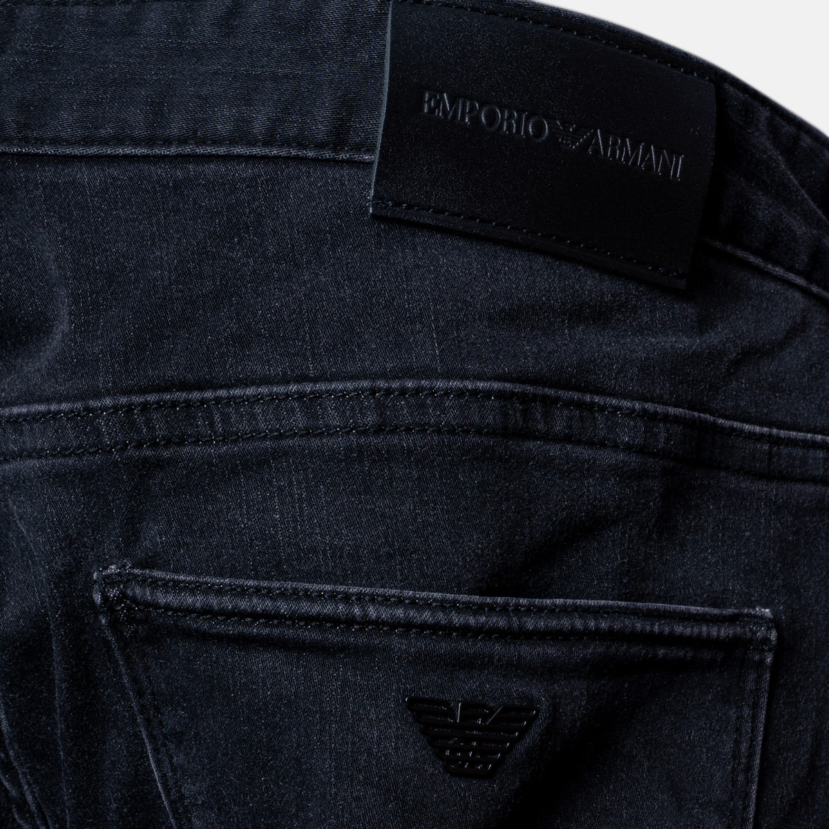 Close-up of the back waistband and pocket area of dark washed denim jeans, showing an embroidered black leather patch with the 'EMPORIO ARMANI' brand name and the signature eagle logo. Emporio Armani Jeans. Sakks Menswear Southampton.
