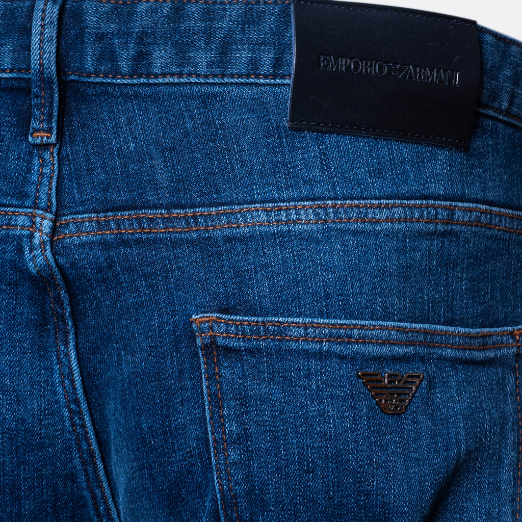Close-up of the back waistband and pocket area of blue denim jeans, showing a black leather patch with the text 'EMPORIO ARMANI' and a small metallic eagle logo on the pocket. Emporio Armani Jeans. Sakks Menswear Southampton.