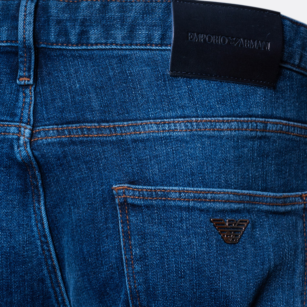 Close-up of the back waistband and pocket area of blue denim jeans, showing a black leather patch with the text 'EMPORIO ARMANI' and a small metallic eagle logo on the pocket. Emporio Armani Jeans. Sakks Menswear Southampton.