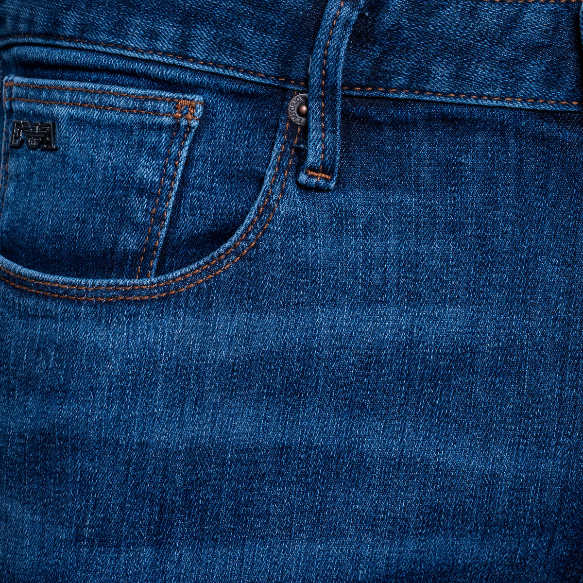 Close-up of the back pocket and waistband of blue denim jeans, showing stitching, a metal rivet, and a small black brand patch on the pocket. Emporio Armani Jeans. Sakks Menswear Southampton.