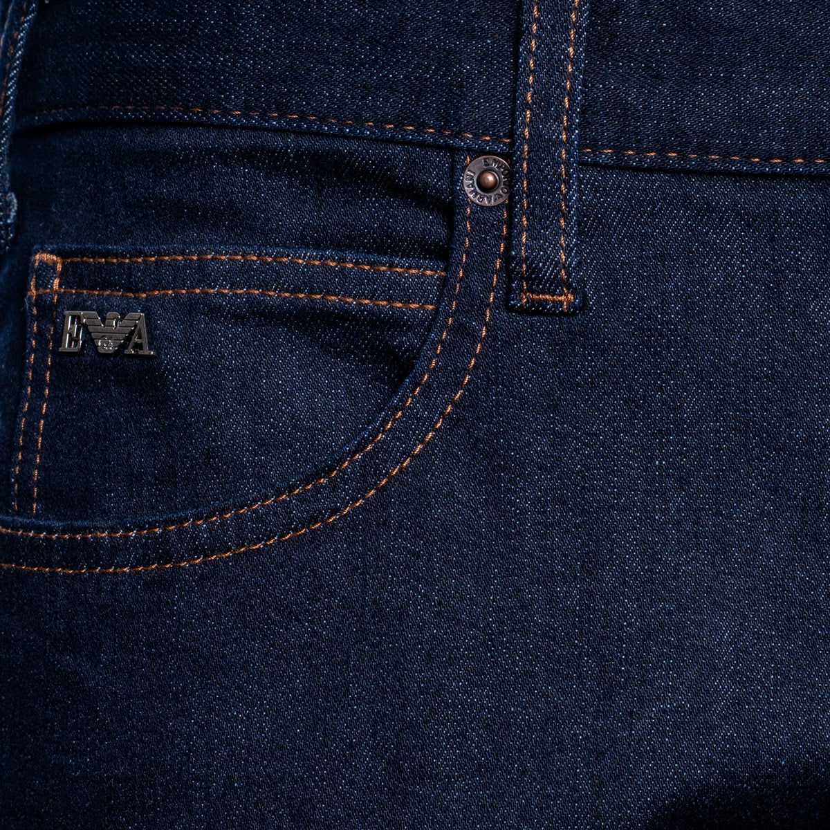 Close-up of dark indigo denim jeans showing the back pocket with orange contrast stitching, a metal rivet, and a small metal logo patch. Emporio Armani Jeans. Sakks Menswear Southampton.