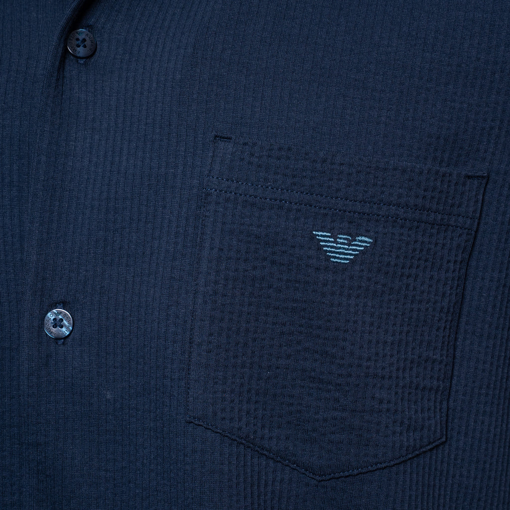 Close-up of a dark navy textured button-up shirt with a chest pocket featuring a light blue embroidered eagle logo, showing part of the button closure. Emporio Armani Short Sleeve Shirt. Sakks Menswear Southampton.