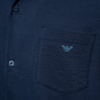 Close-up of a dark navy textured button-up shirt with a chest pocket featuring a light blue embroidered eagle logo, showing part of the button closure. Emporio Armani Short Sleeve Shirt. Sakks Menswear Southampton.