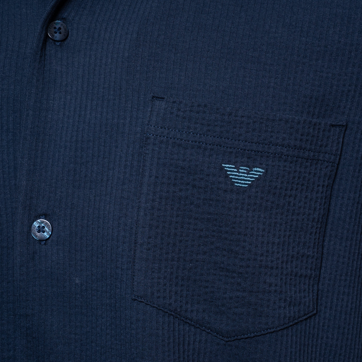 Close-up of a dark navy textured button-up shirt with a chest pocket featuring a light blue embroidered eagle logo, showing part of the button closure. Emporio Armani Short Sleeve Shirt. Sakks Menswear Southampton.