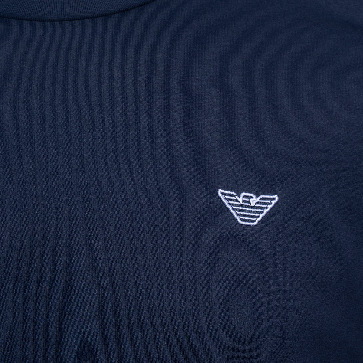Close-up of a navy blue crewneck sweatshirt featuring the white embroidered logo of Emporio Armani on the chest. Emporio Armani T-Shirt. Sakks Menswear Southampton.