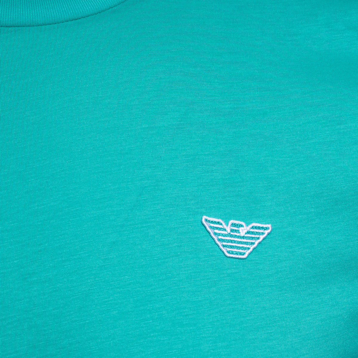 Close-up of a turquoise knit fabric with a small white embroidered Emporio Armani eagle logo on the chest. Emporio Armani T-Shirt. Sakks Menswear Southampton.