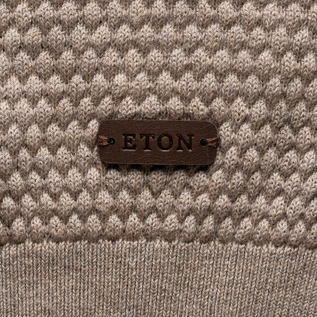 Close-up of a textured knit sweater in taupe and cream with a dark brown leather patch on the chest reading 'ETON'. Eton Knitwear available at Sakks Menswear in Southampton.
