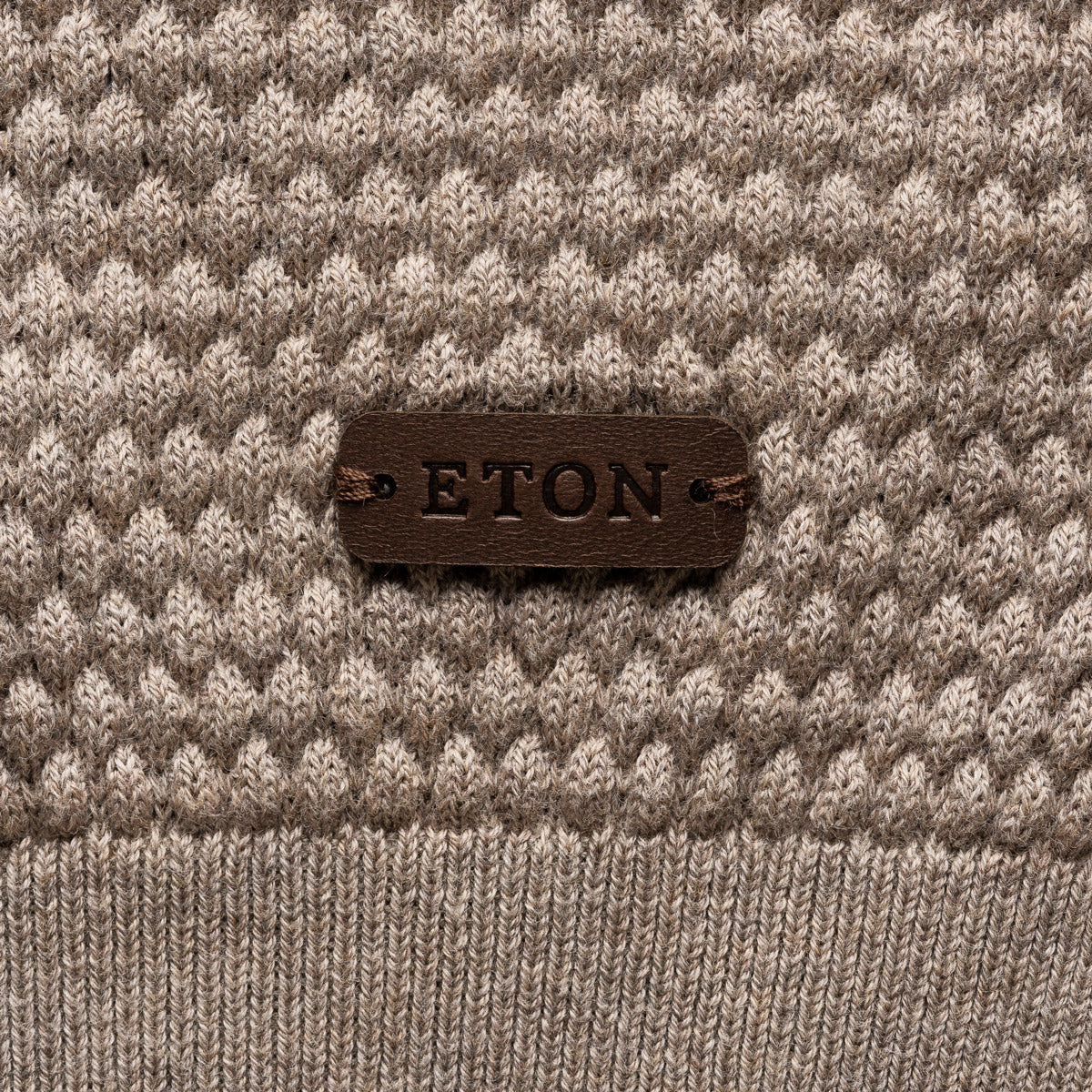 Close-up of a textured knit sweater in taupe and cream with a dark brown leather patch on the chest reading 'ETON'. Eton Knitwear available at Sakks Menswear in Southampton.