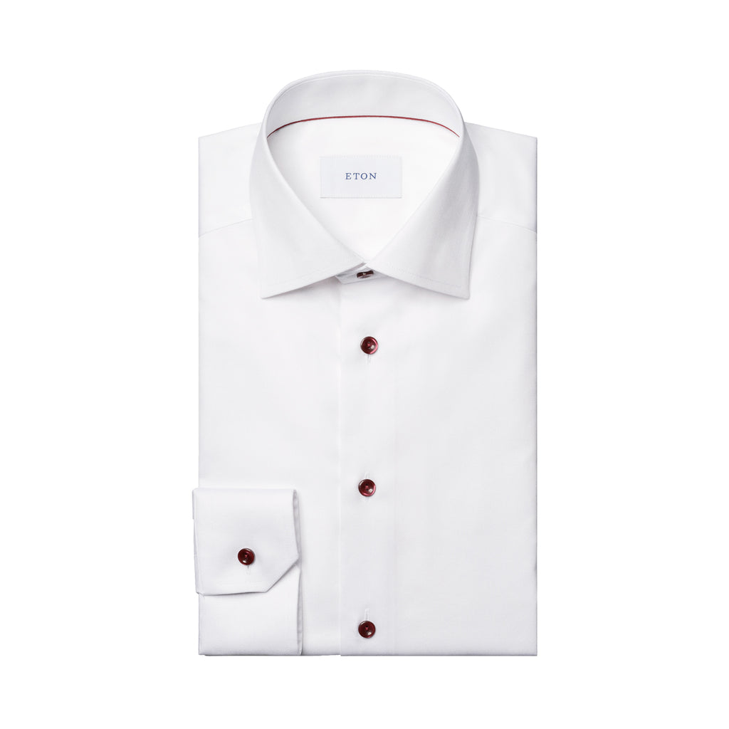 Folded up white shirt with burgundy buttons and detail inside the collar. The tag on the inside of the shirt reads "ETON". The shirt is placed in front of a crisp white background. Eton shirt available at Sakks Menswear in Southampton.