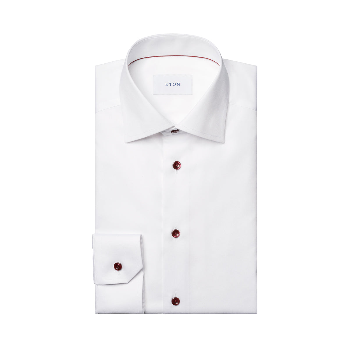 Folded up white shirt with burgundy buttons and detail inside the collar. The tag on the inside of the shirt reads "ETON". The shirt is placed in front of a crisp white background. Eton shirt available at Sakks Menswear in Southampton.