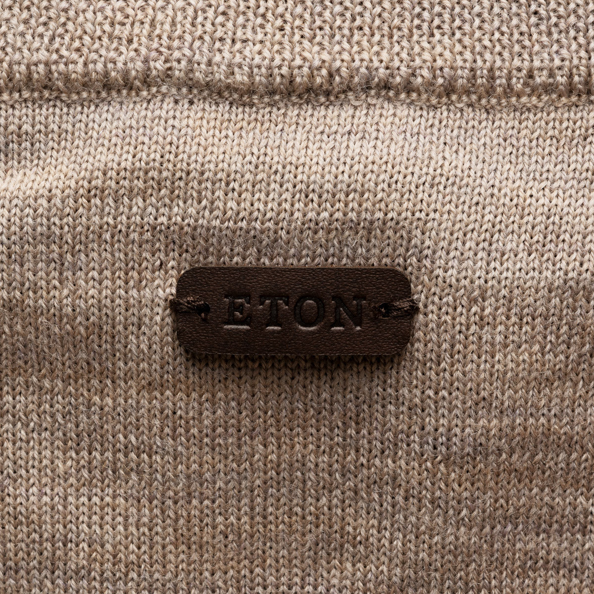 Close-up of a beige knit sweater with a dark brown leather patch on the back reading 'ETON' in raised letters. Eton Cardigan available at Sakks Menswear in Southampton.